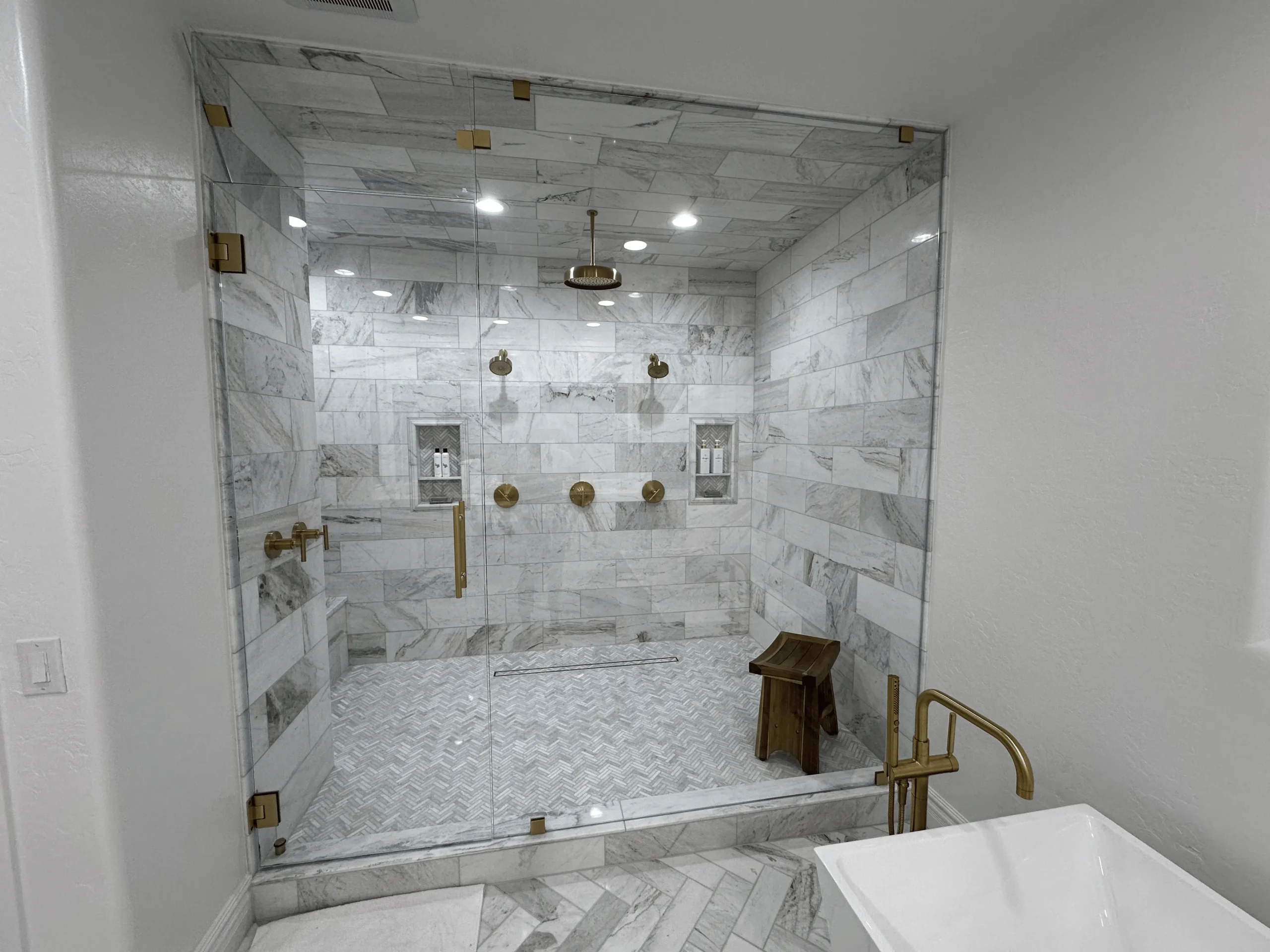 Frameless glass shower enclosure with brass hardware, marble tile walls, and dual shower fixtures in a luxury Arizona bathroom, installed by Clear Cut Glass and Tint