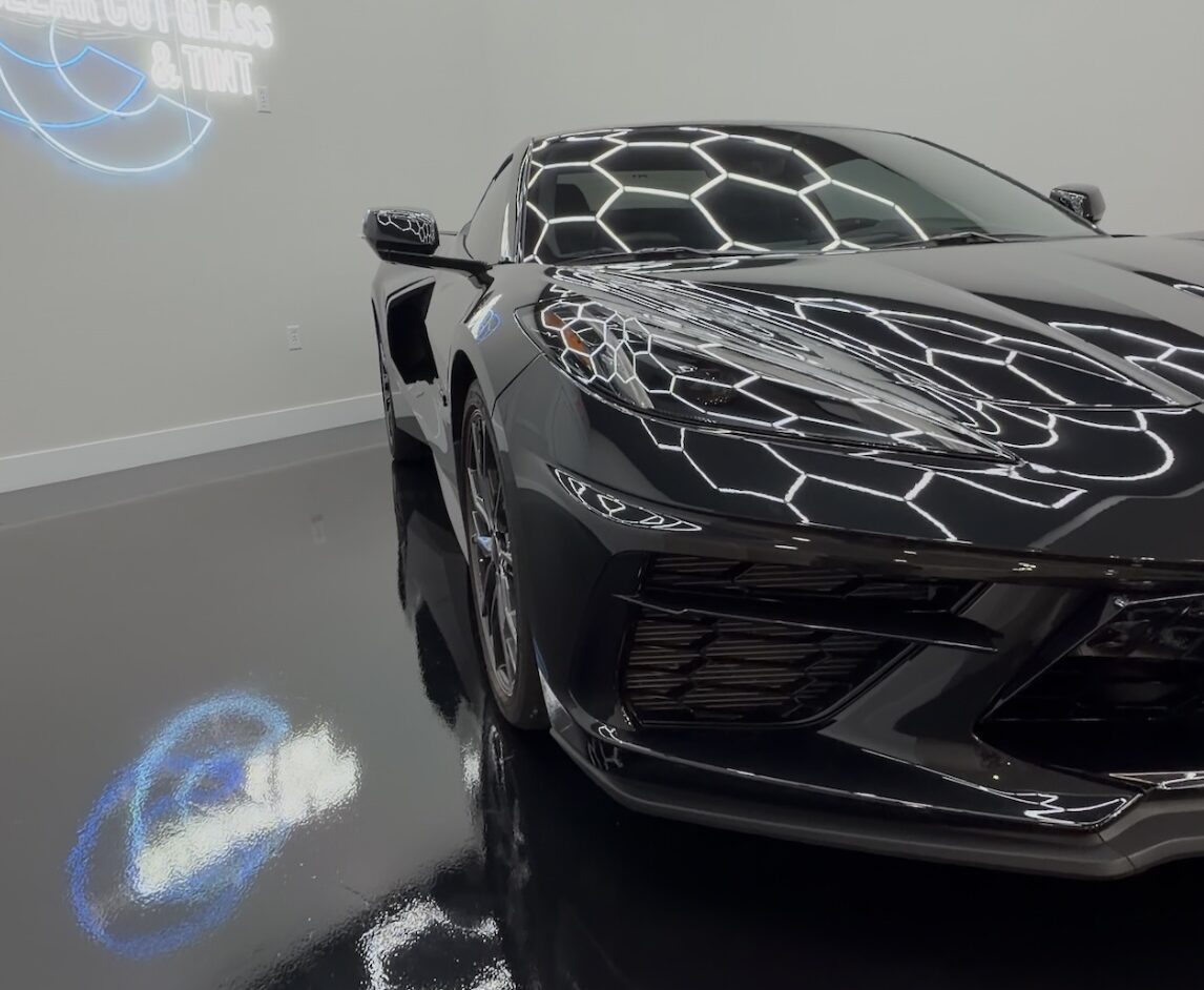 Front three-quarter view of a black Chevrolet Corvette with glossy paint reflecting hexagonal lights at Clear Cut Glass & Tint.