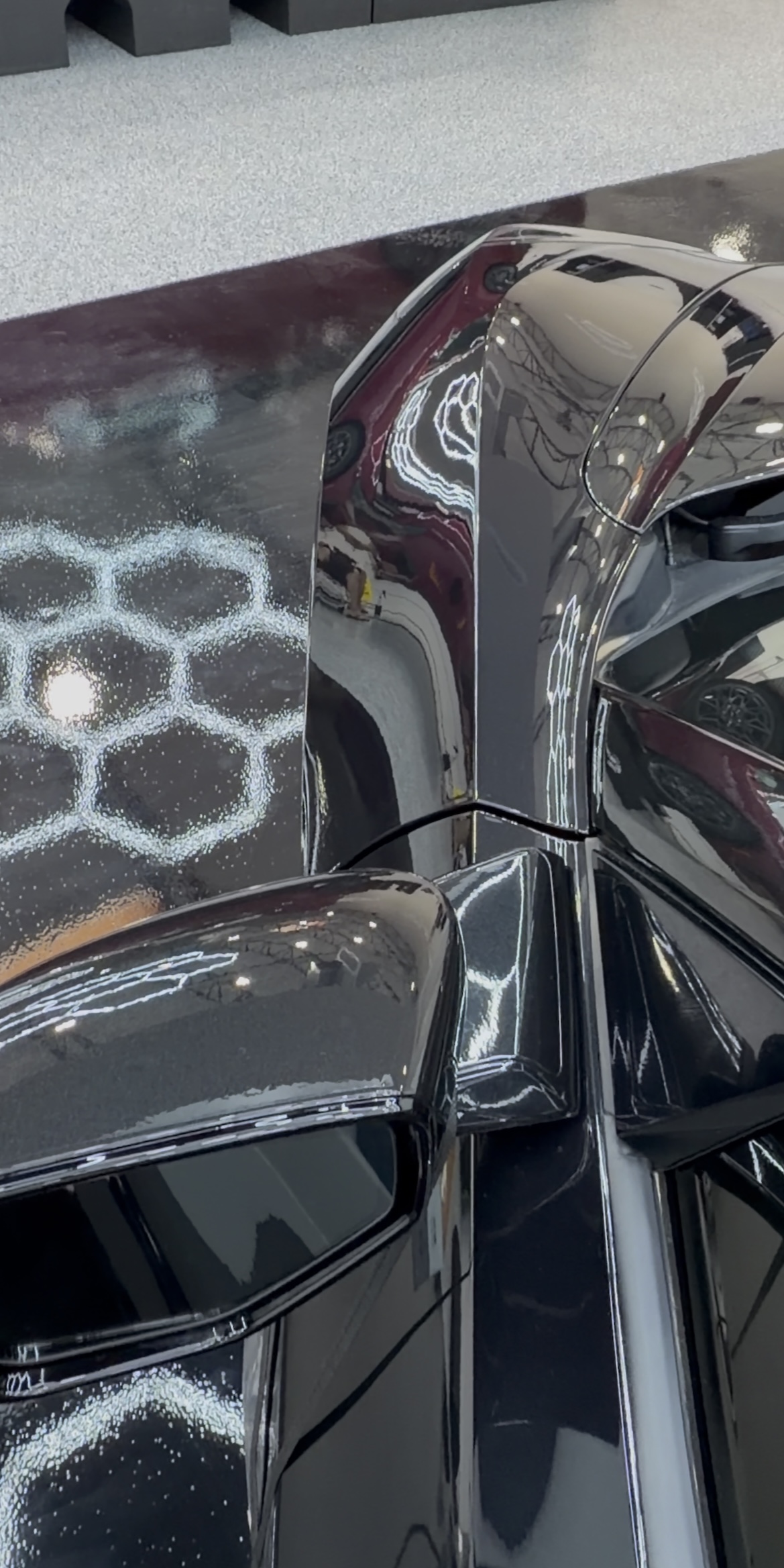 Overhead close-up of a black Chevrolet Corvette showing glossy body panels and mirror-like reflections of shop lighting.